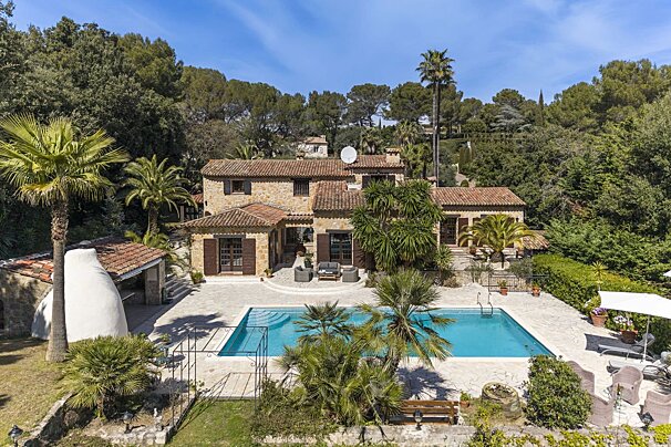 Aerial view of a rustic stone villa with a pool, surrounded by palm trees and lush green hills under a clear sky.