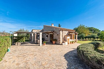A charming stone finca with a paved courtyard, a covered pergola, and green landscaping under a clear blue sky on a sunny day.