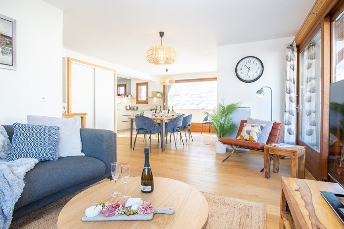 Cozy, modern living space with wooden floors, a grey sofa, coffee table with wine, and an open dining area in the background.