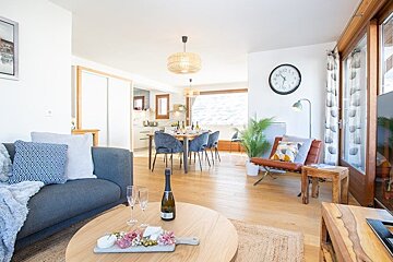 Cozy, modern living space with wooden floors, a grey sofa, coffee table with wine, and an open dining area in the background.