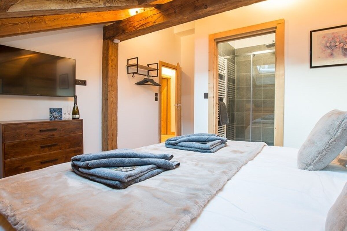 Cozy bedroom with exposed wooden beams, a bed with folded grey towels, and an open doorway showing a modern glass-walled shower.