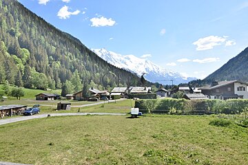 Alpine village nestled in a green valley, surrounded by pine forests and majestic, snow-capped mountains under a blue sky.