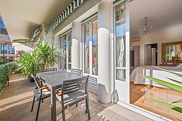 A sunny balcony with outdoor dining furniture, potted plants, and a striped awning, overlooking a luxurious bedroom with a large bed and chandelier.