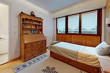 A cozy, rustic bedroom with a wooden desk-cabinet, single bed, and a large window. A snowflake-patterned rug sits on the wooden floor.