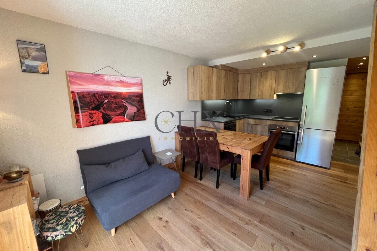 Cozy apartment interior featuring a grey sofa, a wooden dining table, and a kitchen with wooden cabinets and stainless steel appliances.