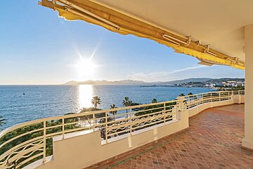 A sunlit balcony with a yellow awning overlooks a sparkling blue ocean at sunset, showing a distant coast, palm trees, and a tiled floor.