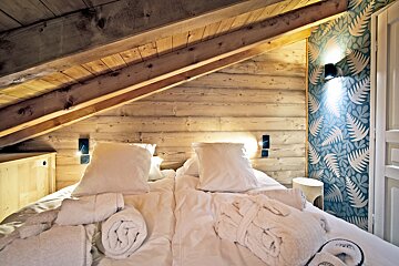 Cozy attic bedroom with sloped wooden beams, a bed with white linens and towels, and a blue fern-patterned accent wall.