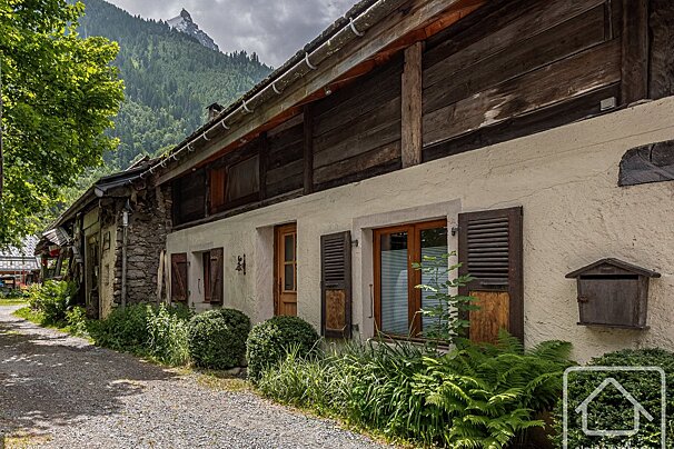 An old Alpine house with a gravel path and lush plants. Tall green mountains rise behind under a cloudy sky.