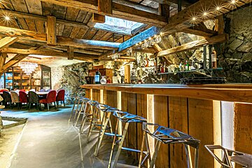 Rustic stone cellar bar with a wooden counter, modern silver stools, and a dining area in the background under exposed beams.