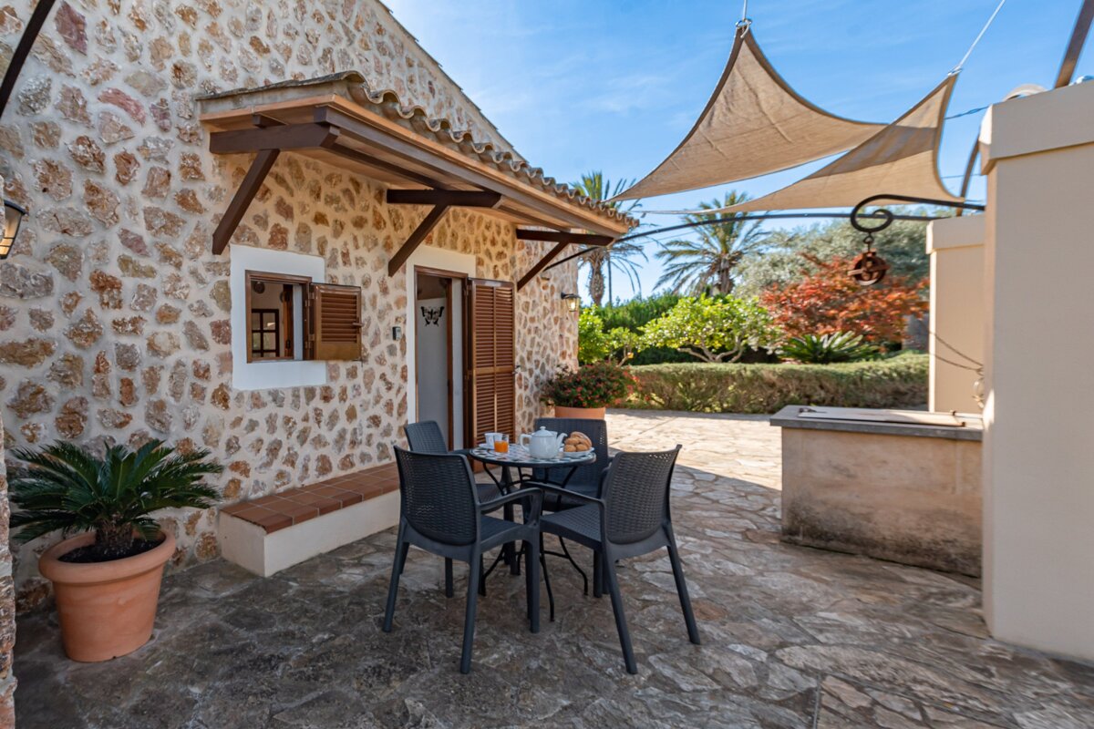 A sunny rustic stone house patio with breakfast on a table for two, surrounded by a lush garden and shaded by sun sails.