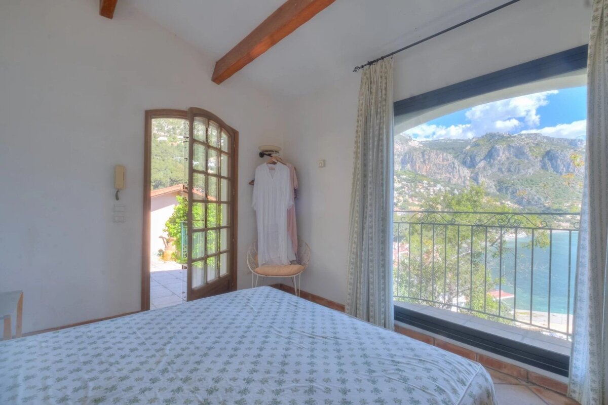 Cozy bedroom with a patterned bedspread, an open door to a sunny patio, and a large window overlooking a scenic mountain and sea landscape.