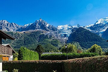 Majestic snow-capped mountains and glaciers tower over lush green forests, with hedges and a building in the sunny foreground under a clear blue sky.
