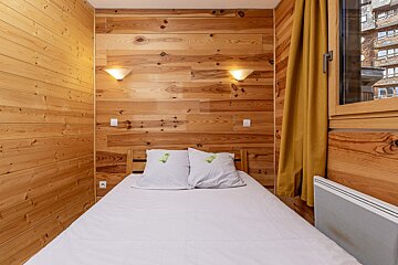 A rustic bedroom with light wood paneling, a bed with white linens and patterned pillows, two wall sconces, and a yellow curtain next to a window.