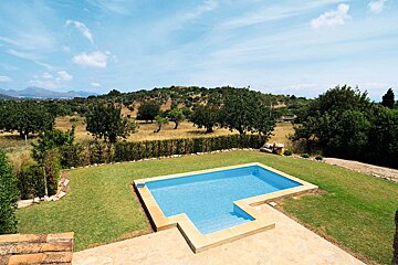 An inviting outdoor swimming pool in a green lawn, overlooking a rustic landscape of dry fields, trees, and distant hills under a bright blue sky.