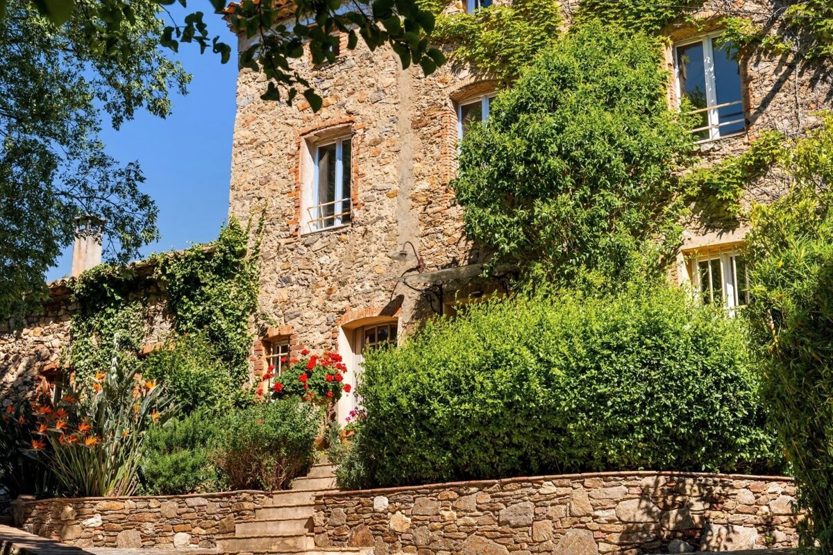 A sunlit, vine-covered stone house with vibrant red and orange flowers, lush green foliage, and stone steps under a clear blue sky.