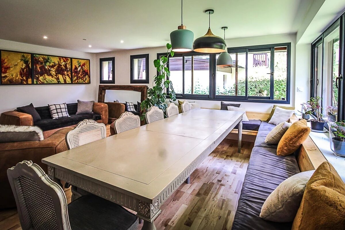 A bright, modern dining and living area features a long table, cane-back chairs, a cozy sofa, and built-in window bench seating with garden views.