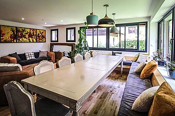 A bright, modern dining and living area features a long table, cane-back chairs, a cozy sofa, and built-in window bench seating with garden views.