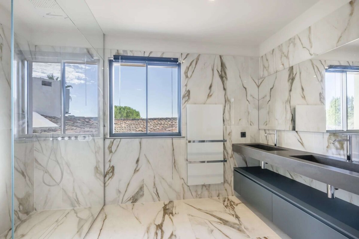 A modern bathroom featuring white marble walls/floor, a large window with rooftop view, a glass shower, and a sleek double vanity with dark sinks.