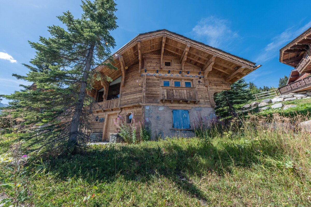 A rustic wooden chalet with intricate details stands on a sunny, grassy hillside, flanked by a tall pine tree under a clear blue sky.