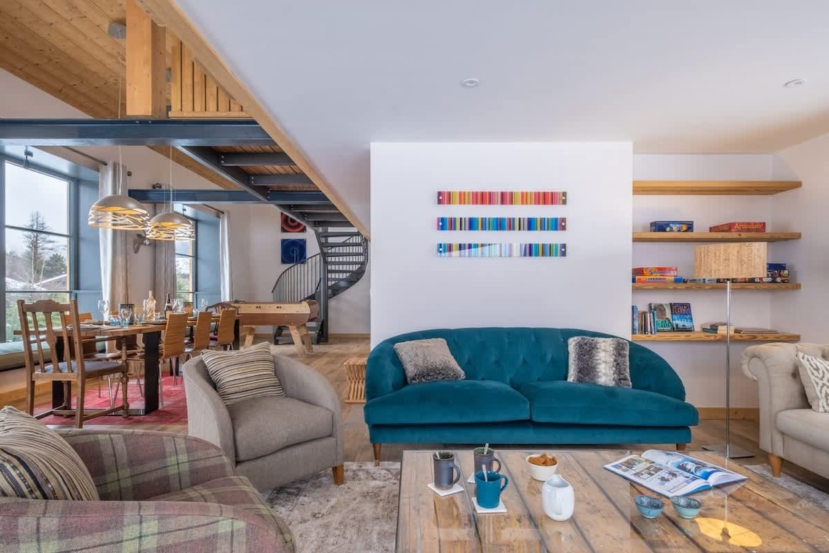 Cozy open-plan living area featuring a teal velvet sofa, armchairs, and a dining table with a spiral staircase in the background.