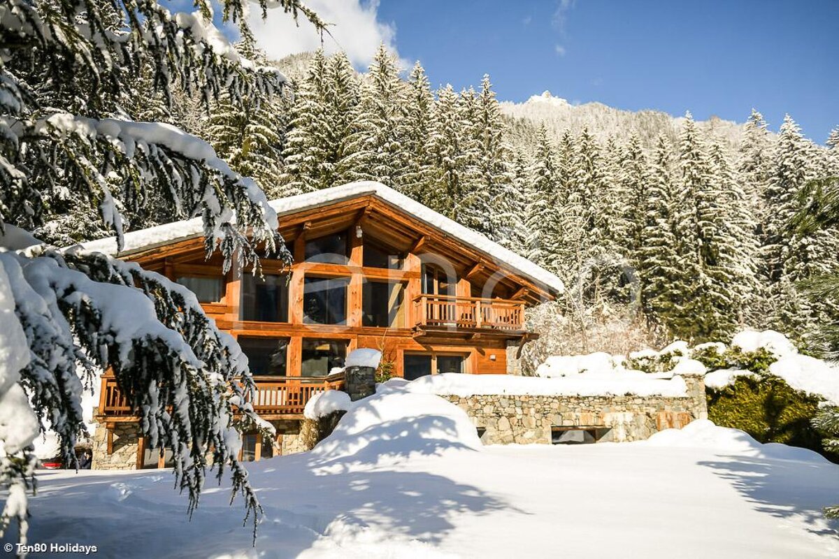 A cozy wooden chalet nestled in a snowy mountain forest with snow-covered pine trees under a bright blue sky.