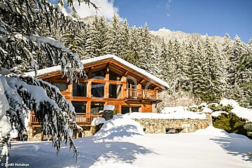 A cozy wooden chalet nestled in a snowy mountain forest with snow-covered pine trees under a bright blue sky.