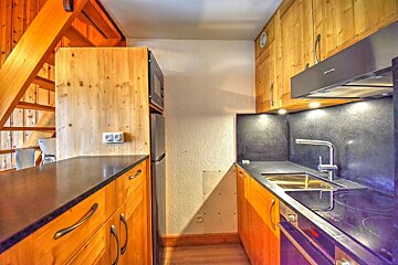 A compact kitchen features light wood cabinets, dark countertops, modern appliances (sink, stove, oven, microwave, fridge), and a wooden staircase.