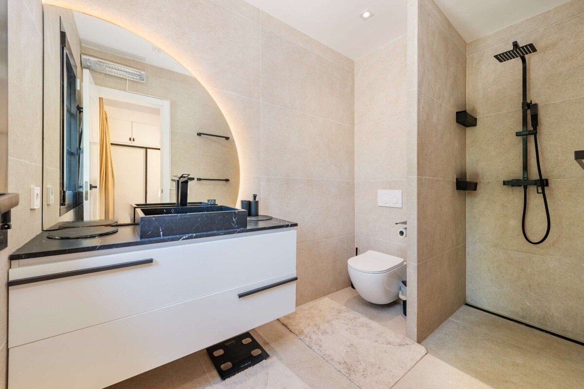 A modern bathroom with beige tiles, a black marble sink on a white floating vanity, an illuminated mirror, a toilet, and a walk-in shower with black fixtures.