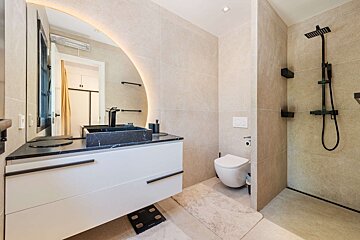 A modern bathroom with beige tiles, a black marble sink on a white floating vanity, an illuminated mirror, a toilet, and a walk-in shower with black fixtures.