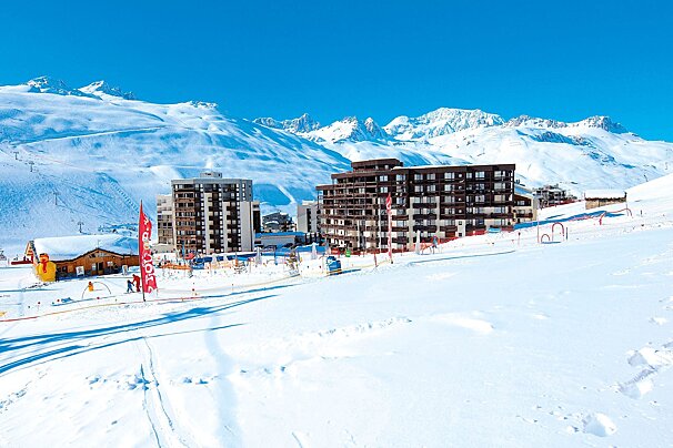 A sunny ski resort with modern buildings nestled amidst vast snow-covered mountains under a clear blue sky, with ski slopes in the foreground.