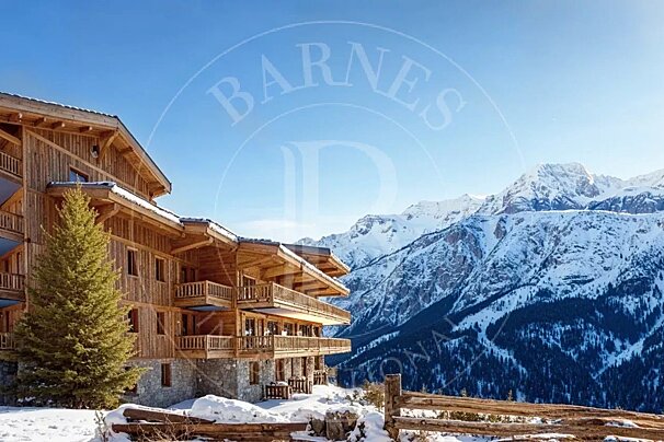 A wooden alpine chalet stands in a snowy landscape with majestic snow-covered mountains under a clear blue sky.
