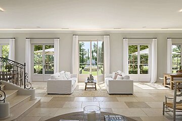 Bright, elegant living room with white sofas, French doors opening to a lush garden, and a grand staircase. Bathed in natural light.