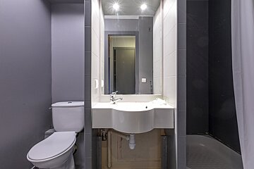 A compact bathroom with a white toilet, sink under an illuminated mirror, and a dark-tiled shower area. Walls are gray with white tile accents.