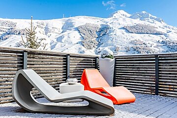 Modern orange and grey loungers on a snowy deck with a stunning view of snow-covered mountains under a bright blue sky.