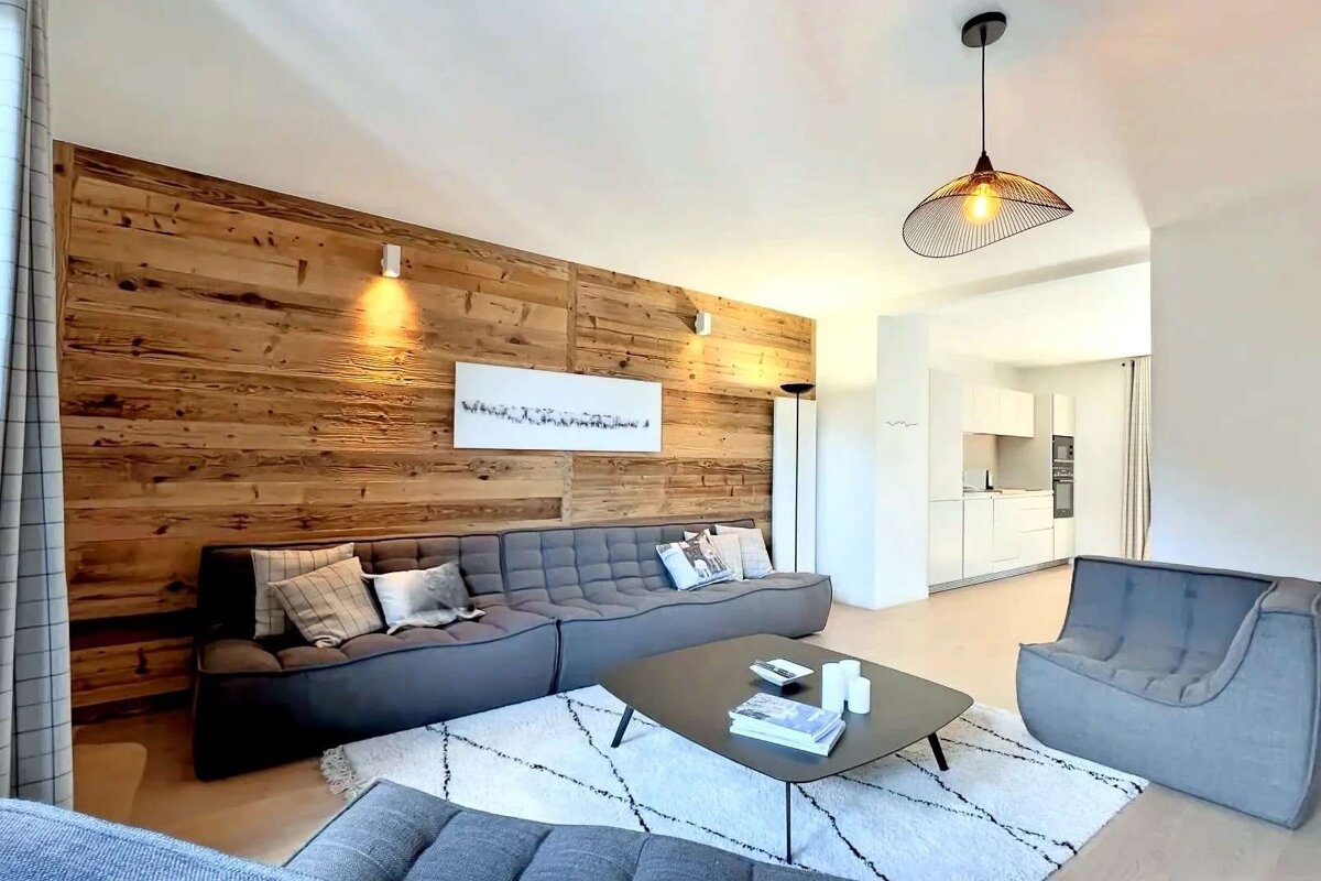 A chic modern living room features a rustic wood accent wall, gray modular sofa, and stylish geometric rug. An open kitchen is visible.