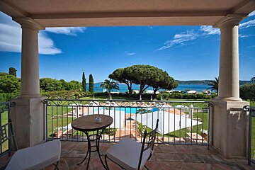 A balcony with a table and chairs offers a scenic view of a lush garden, swimming pool, and the vibrant blue ocean under a clear sky.