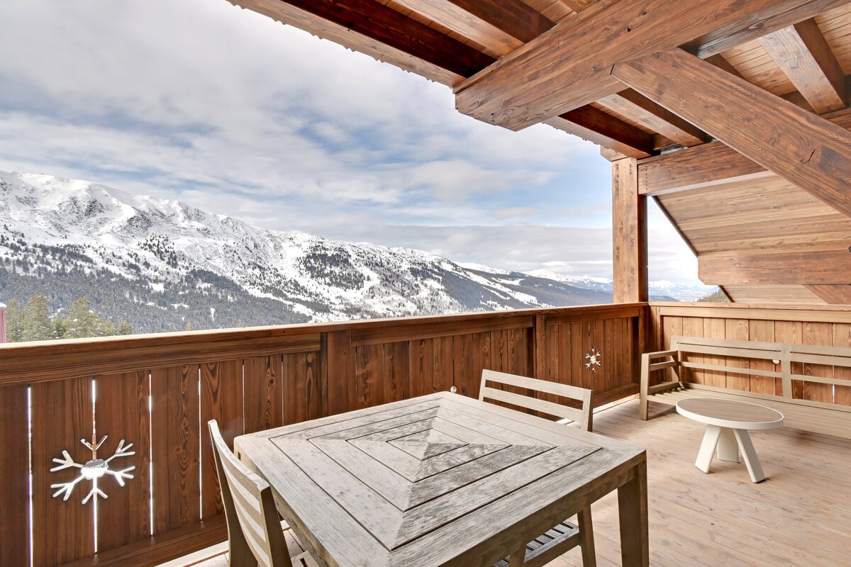 Wooden balcony with table & chairs overlooks snow-capped mountains. Rustic architecture, snowflake decor, cloudy sky.
