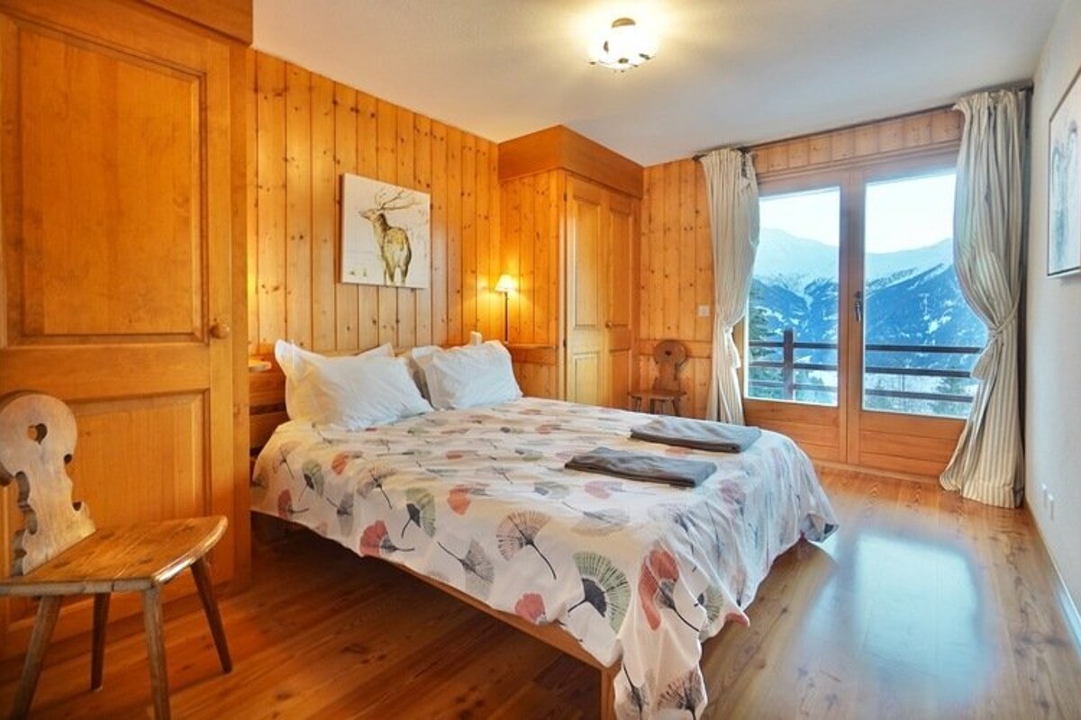 Cozy wood-paneled chalet bedroom with a double bed and a sliding glass door looking out onto a scenic snowy mountain range.