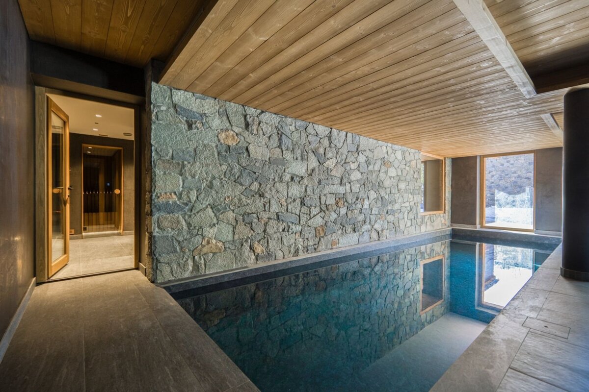 Luxurious indoor swimming pool with a textured stone wall, wooden ceiling, and reflections in the water.