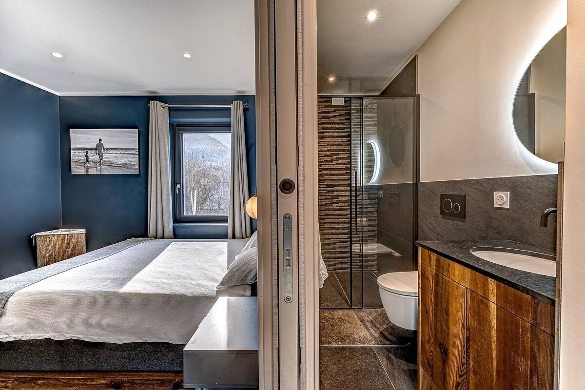 Modern bedroom (dark blue wall, mountain view, bed) next to a sleek bathroom (vanity, backlit mirror, shower, toilet), separated by a sliding door.