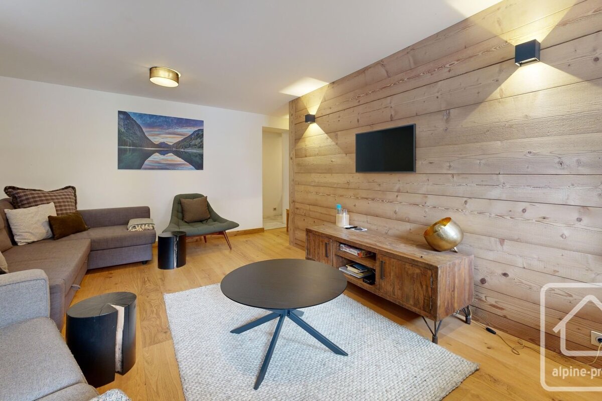 A modern alpine living room featuring a wood-paneled TV wall, sectional sofa, armchair, round coffee table on a rug, and a scenic mountain painting.