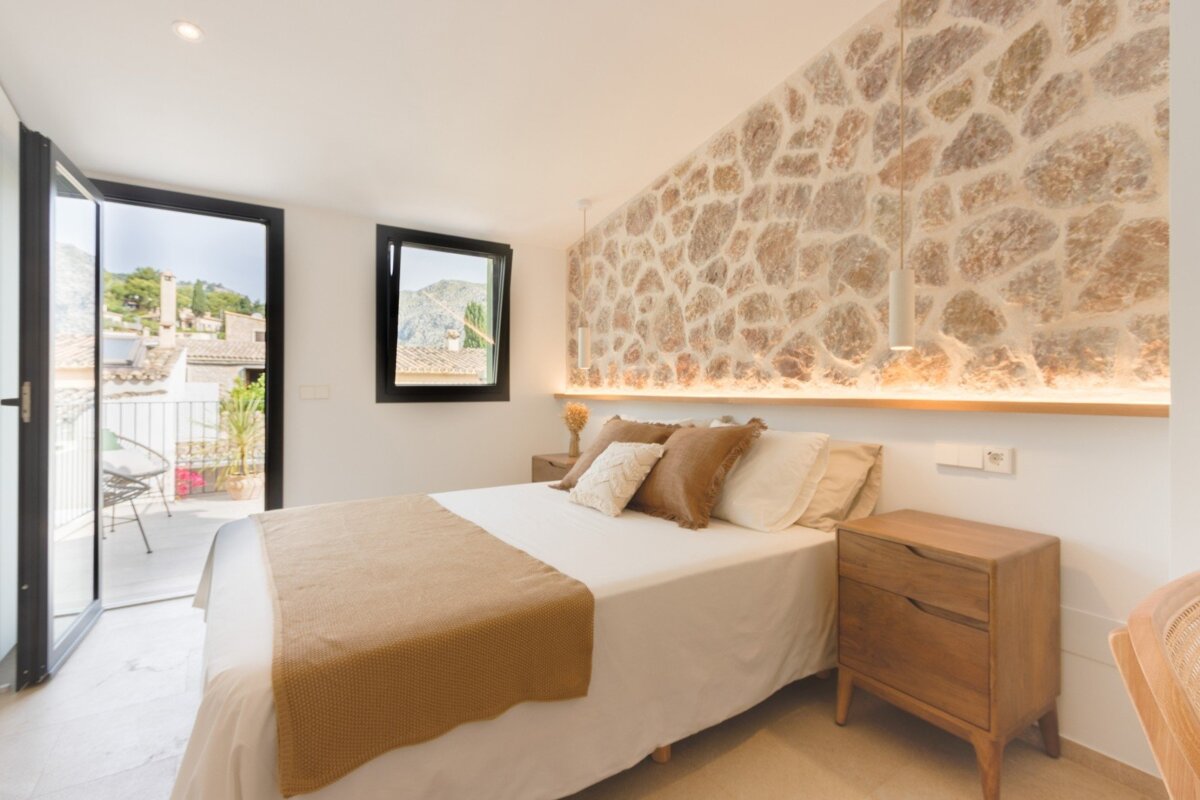 Bright bedroom with a stone accent wall, cozy bed, and a balcony offering a scenic view of a village and mountains.