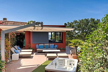A modern outdoor patio features a red building, retractable awning, comfy outdoor seating, and a white hot tub, all set amidst lush greenery under a clear blue sky.