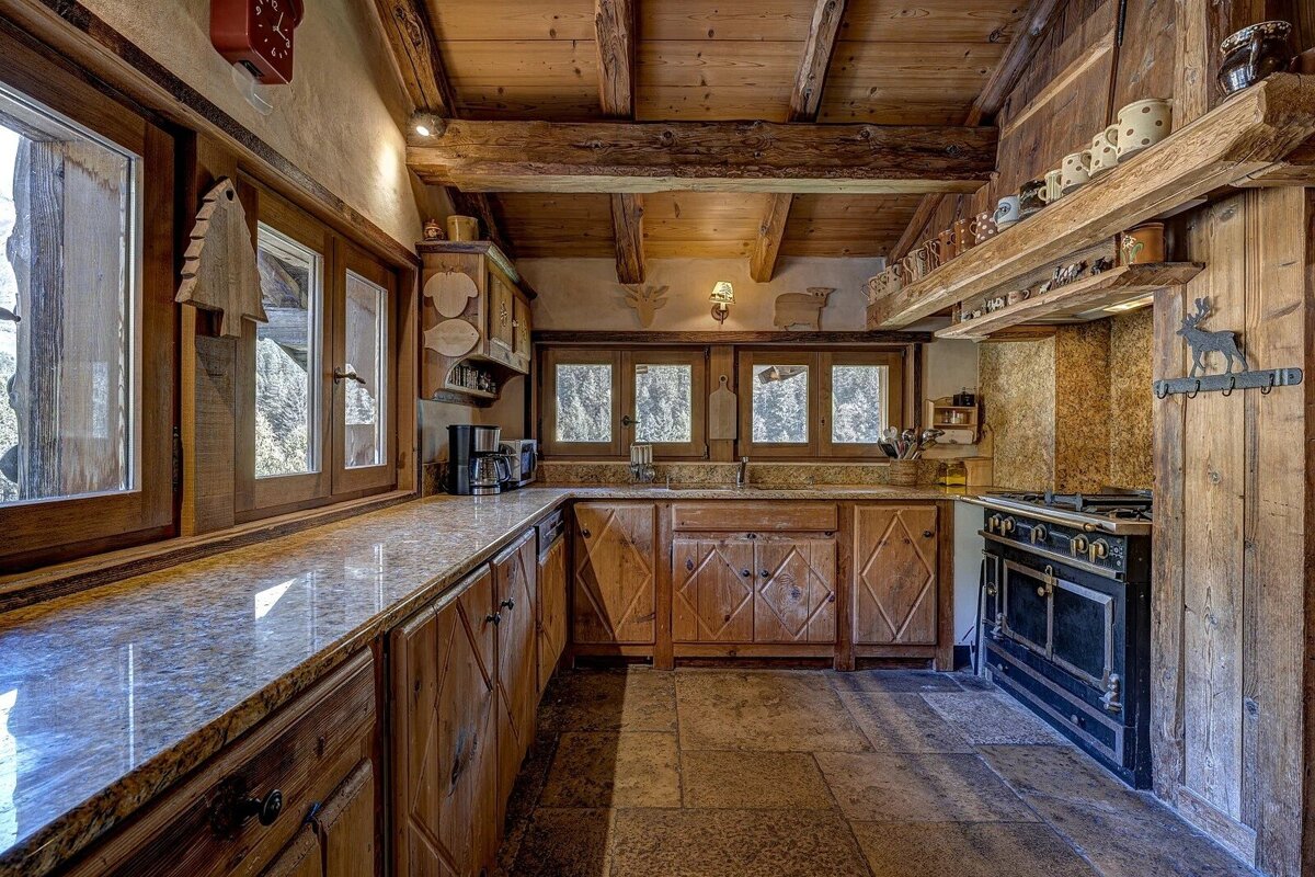 A cozy, rustic wooden kitchen featuring exposed beams, stone countertops, a vintage stove, and large windows with outdoor views.