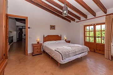 Cozy rustic bedroom with a bed, wooden nightstands, patterned throw, exposed beams, a lantern, tiled floor, and doors opening to a green view.
