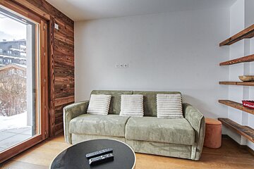 A cozy living room features a green corduroy sofa, wooden accent wall, and a large window showing a snowy landscape. Floating wooden shelves complement the modern rustic design.