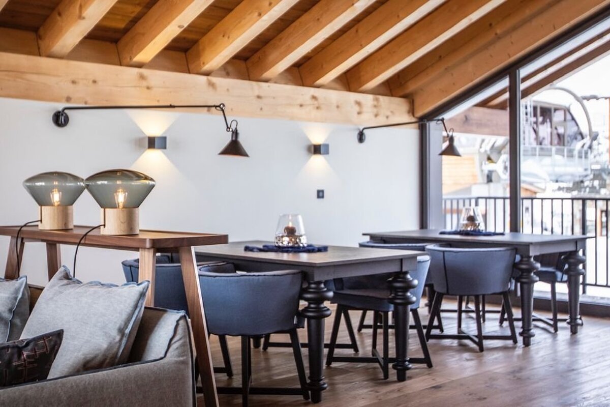 Cozy dining area with exposed wooden ceiling beams, dark tables, blue chairs, and stylish modern lighting.