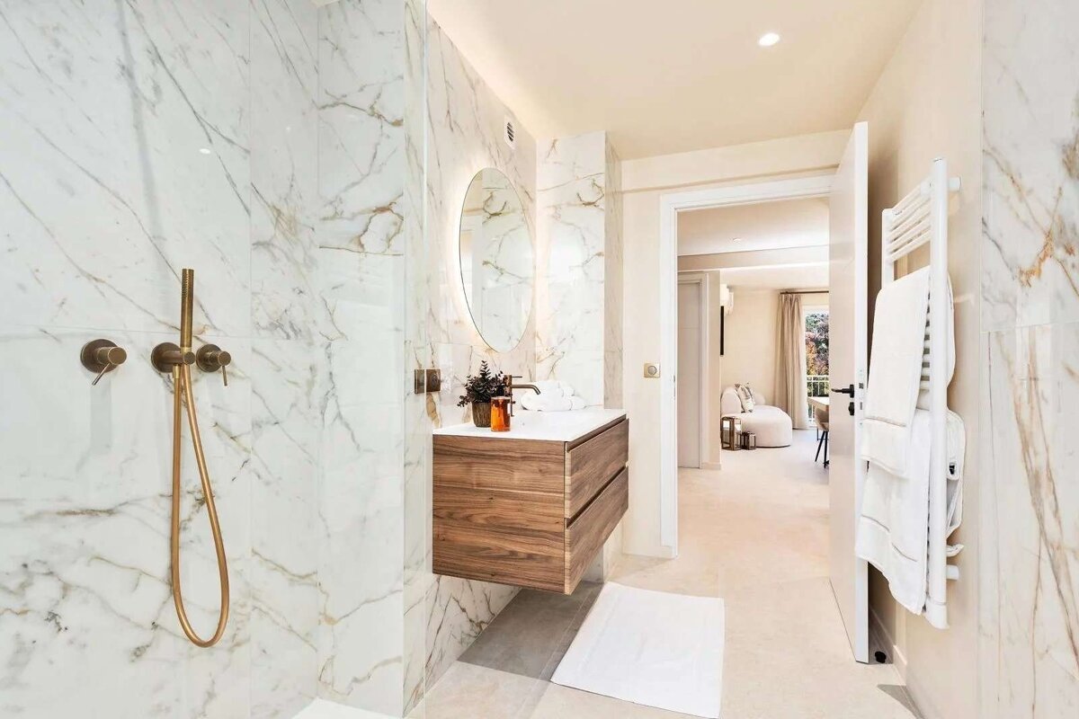 An elegant modern bathroom featuring white marble walls with gold veins, brass fixtures, a floating wooden vanity, and a backlit mirror, opening to a chic room.