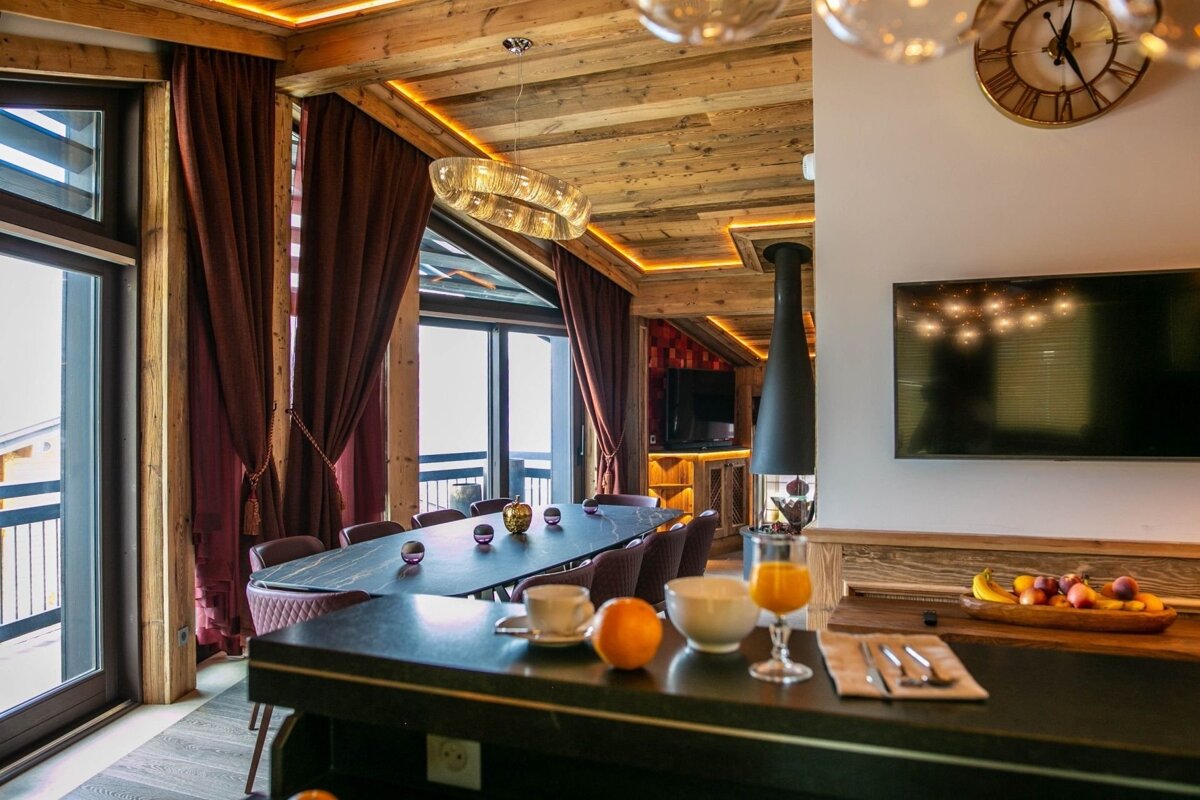Luxurious, warm-lit wood-paneled chalet dining room with a long table, breakfast bar, suspended fireplace, and large windows.