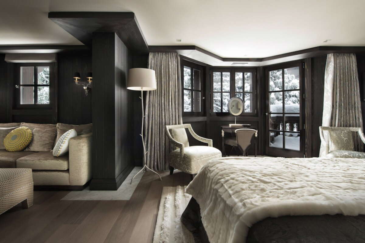 Luxurious dark-paneled bedroom suite with a plush bed, cozy sitting area, and large windows revealing a snowy winter landscape outside.